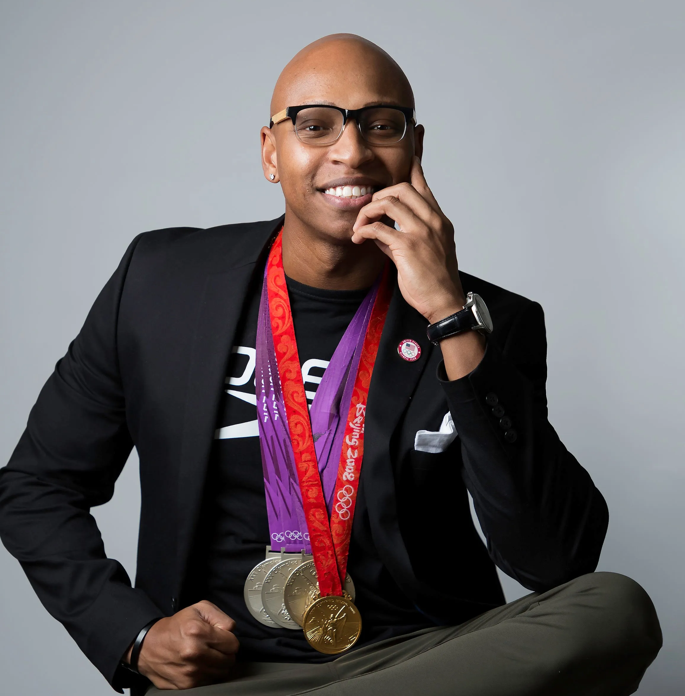 Portrait of a smiling man wearing glasses, a blazer, and Olympic medals around his neck.