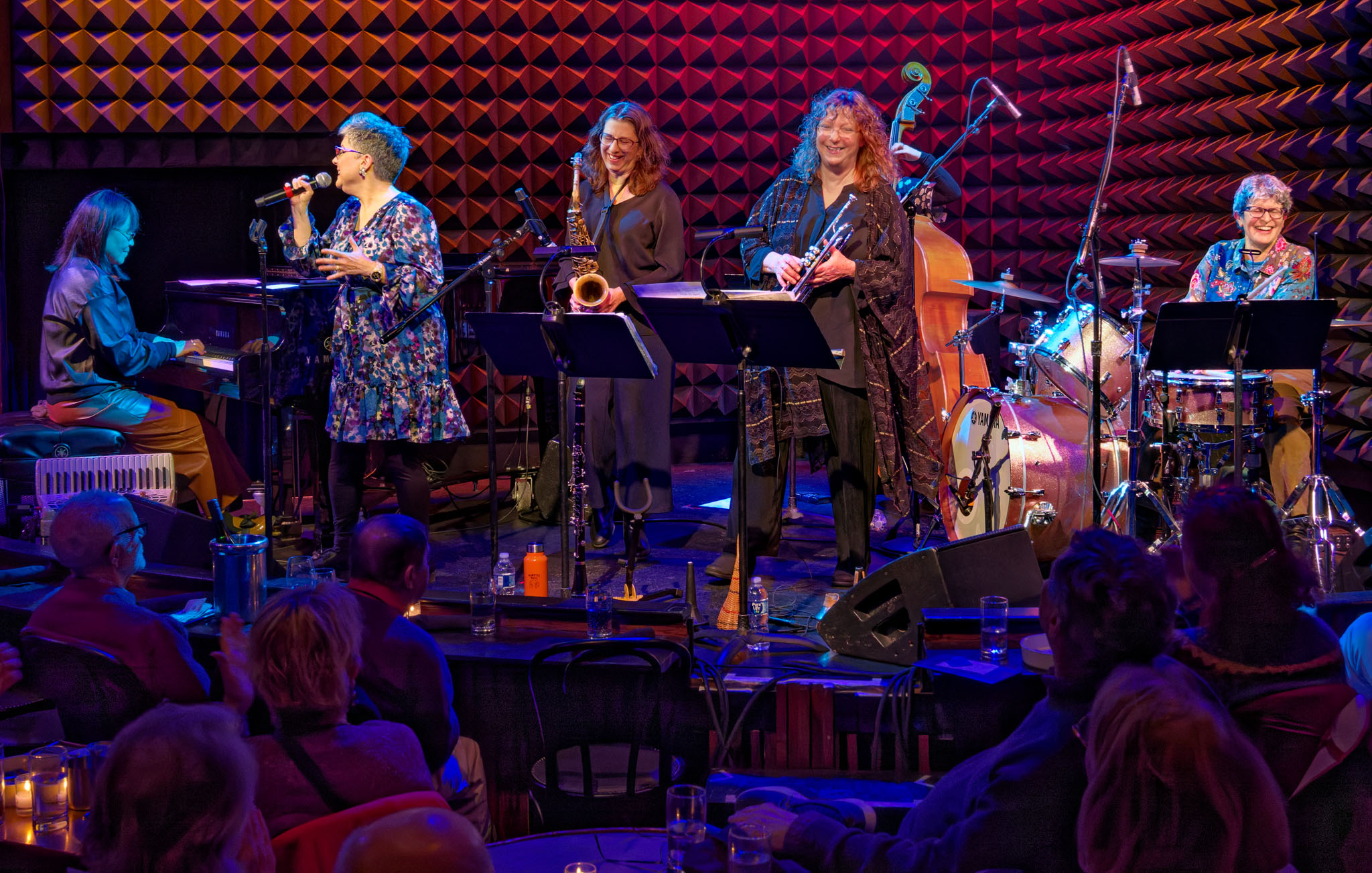 A jazz ensemble performs on stage with a singer, pianist, horn players, bassist, and drummer before a live audience.
