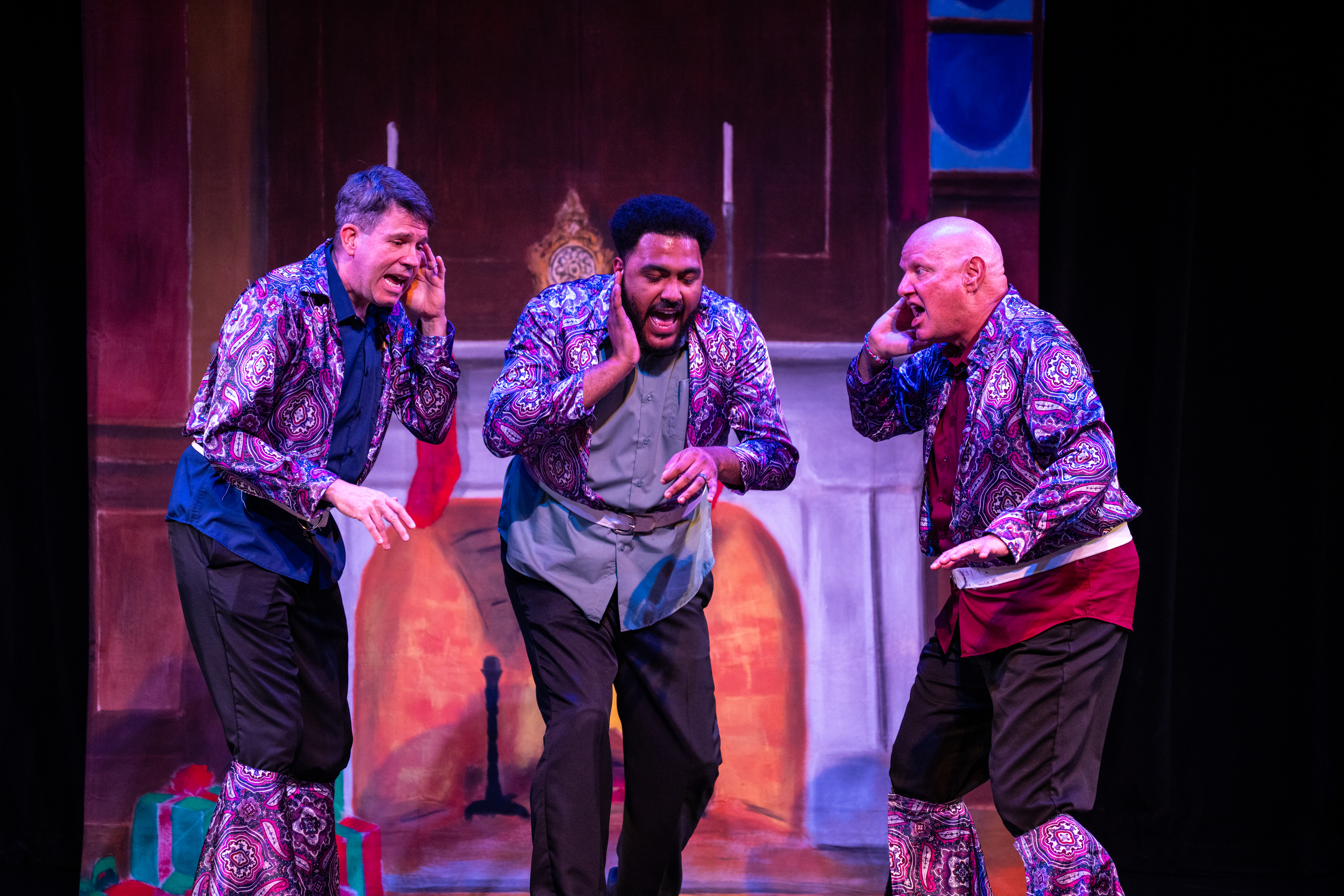 Three performers in colorful costumes sing together on stage in front of a fireplace set.