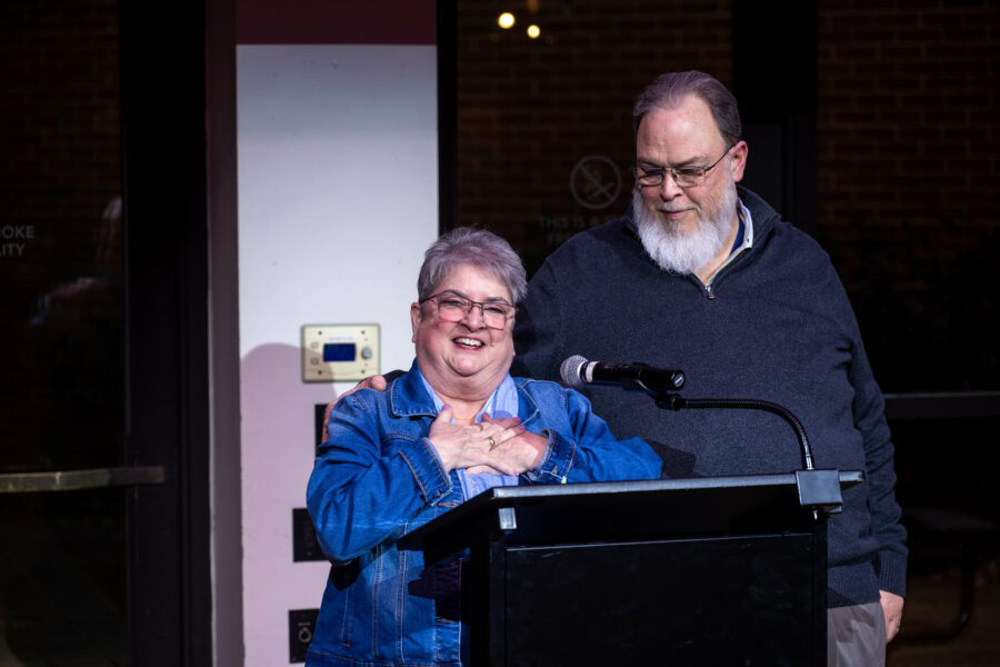 Two people stand together behind a podium with a microphone; the person in front, wearing a denim jacket, has their hands clasped over their chest while the person beside them rests a hand supportively on their shoulder.