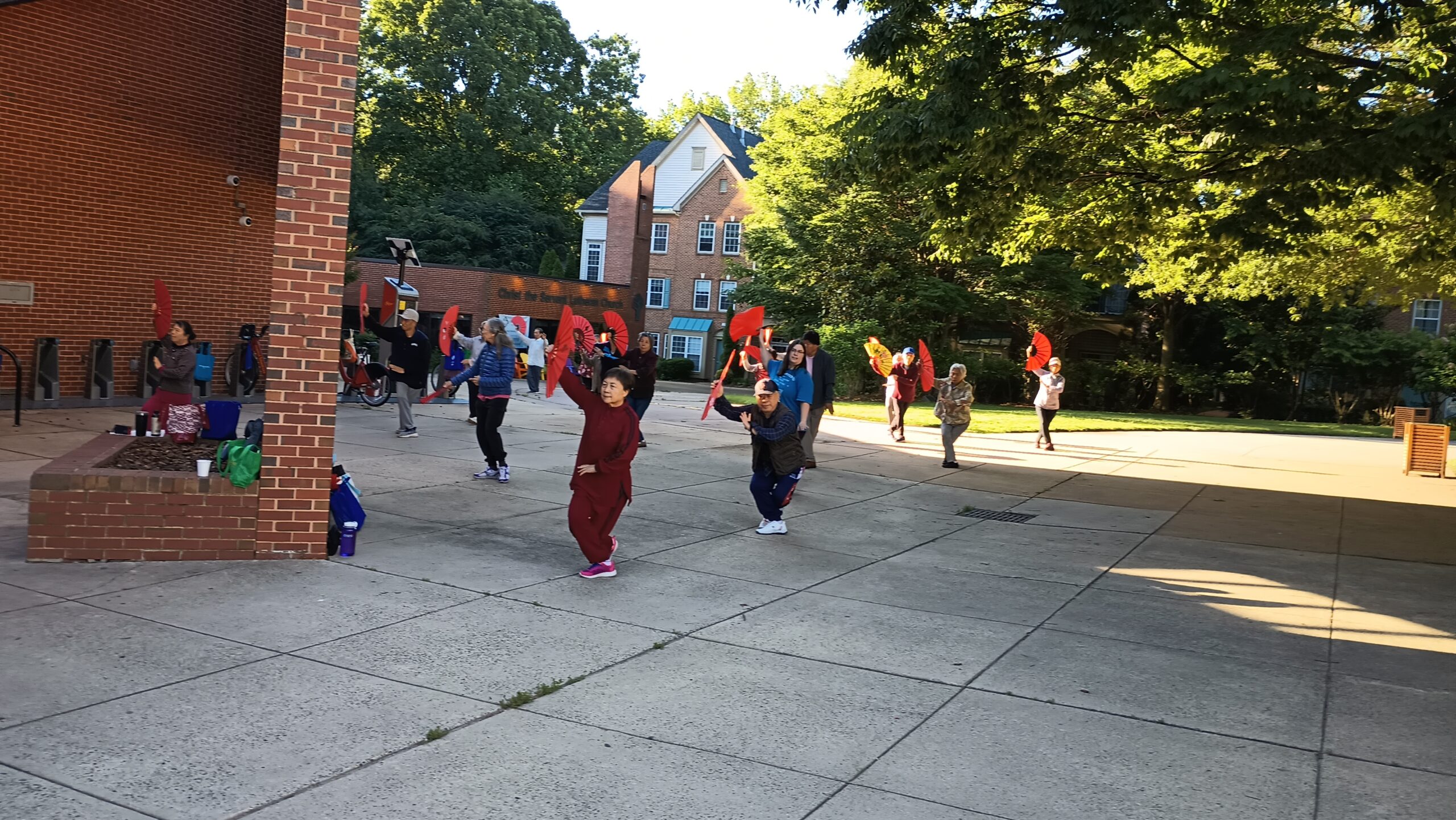 A group of adults practice tai chi outdoors while holding bright red folding fans.