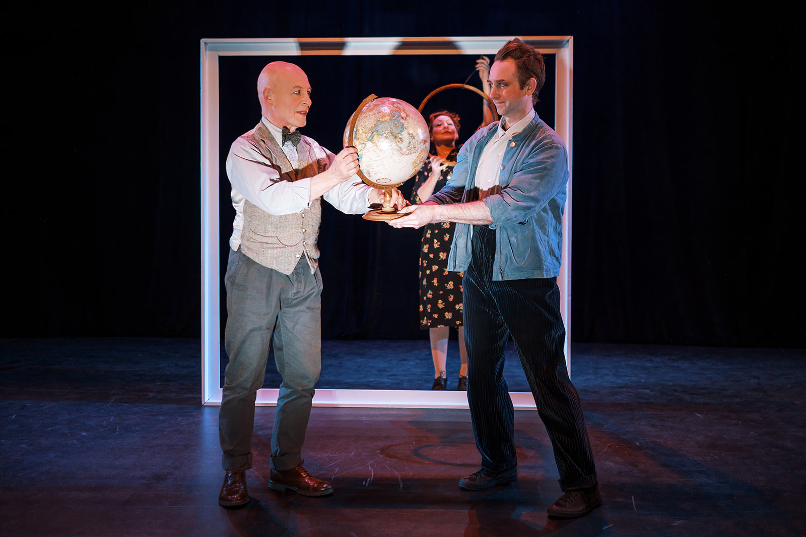 Two performers hold a globe on stage while a third performer stands behind them inside a large rectangular frame.