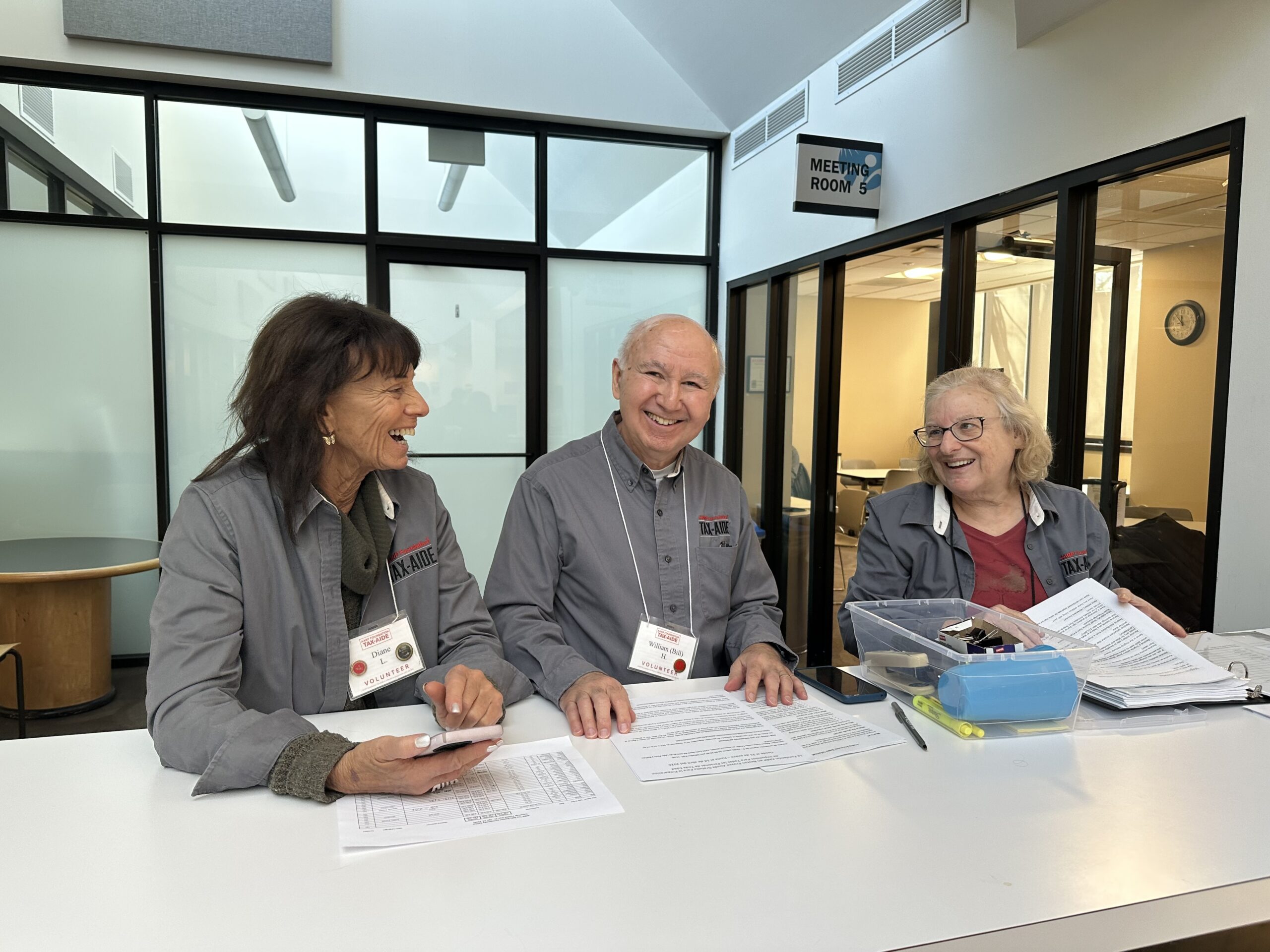 Three older adult volunteers seated at a table reviewing paperwork and smiling during a tax assistance session.
