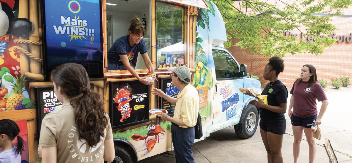 People lined up at a Kona Ice truck receiving shaved ice outdoors.