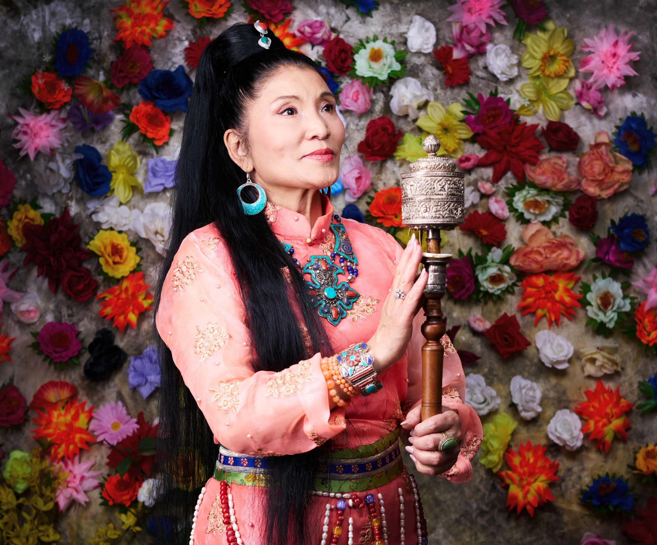 Portrait of a woman in colorful traditional attire holding a prayer wheel in front of a vibrant floral backdrop.