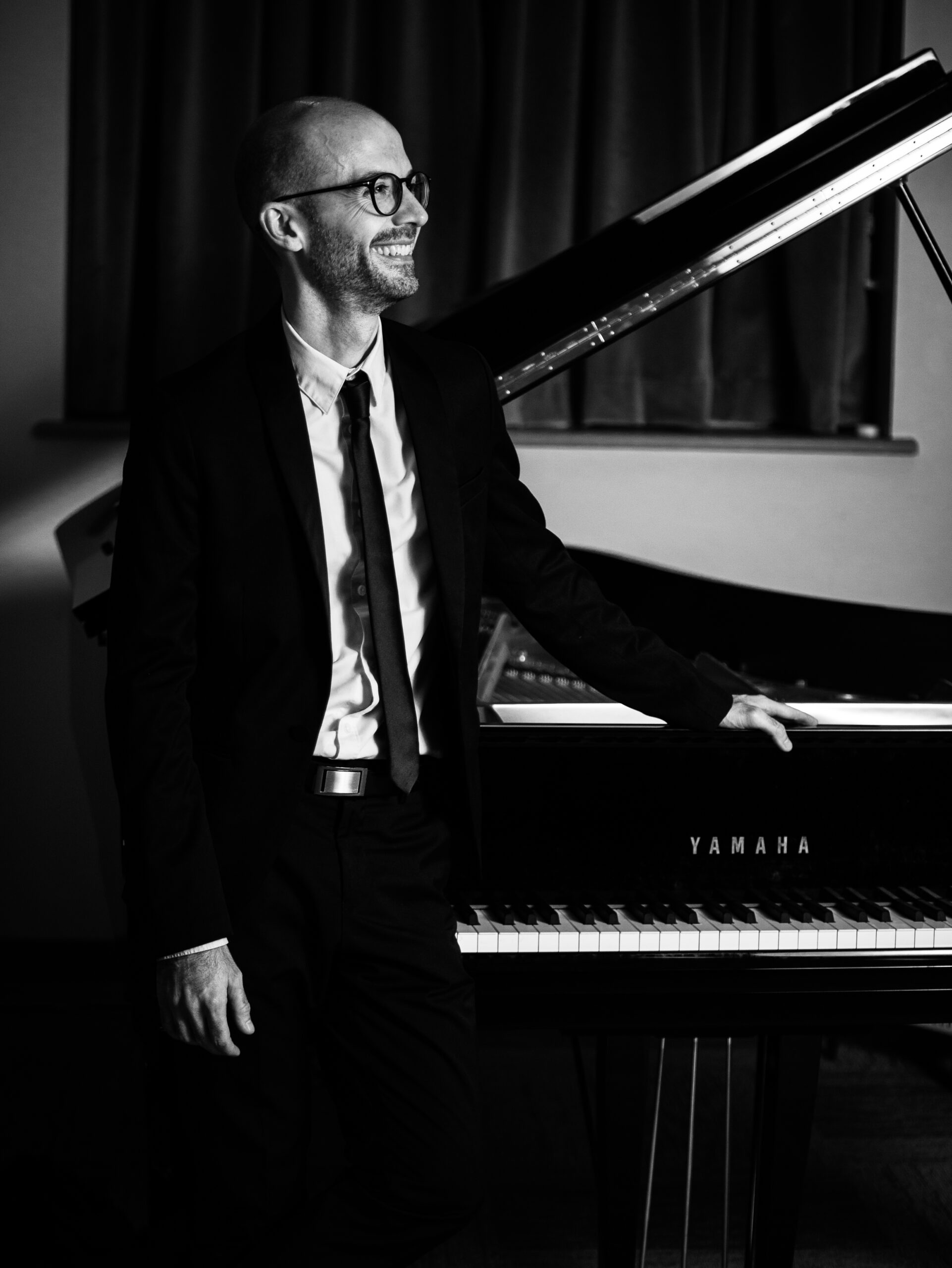 Smiling pianist in a suit standing beside a grand piano.