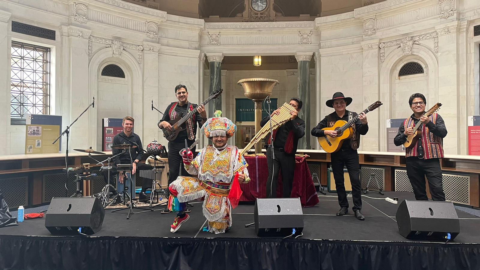 Musicians and a dancer perform traditional Andean music on an indoor stage.