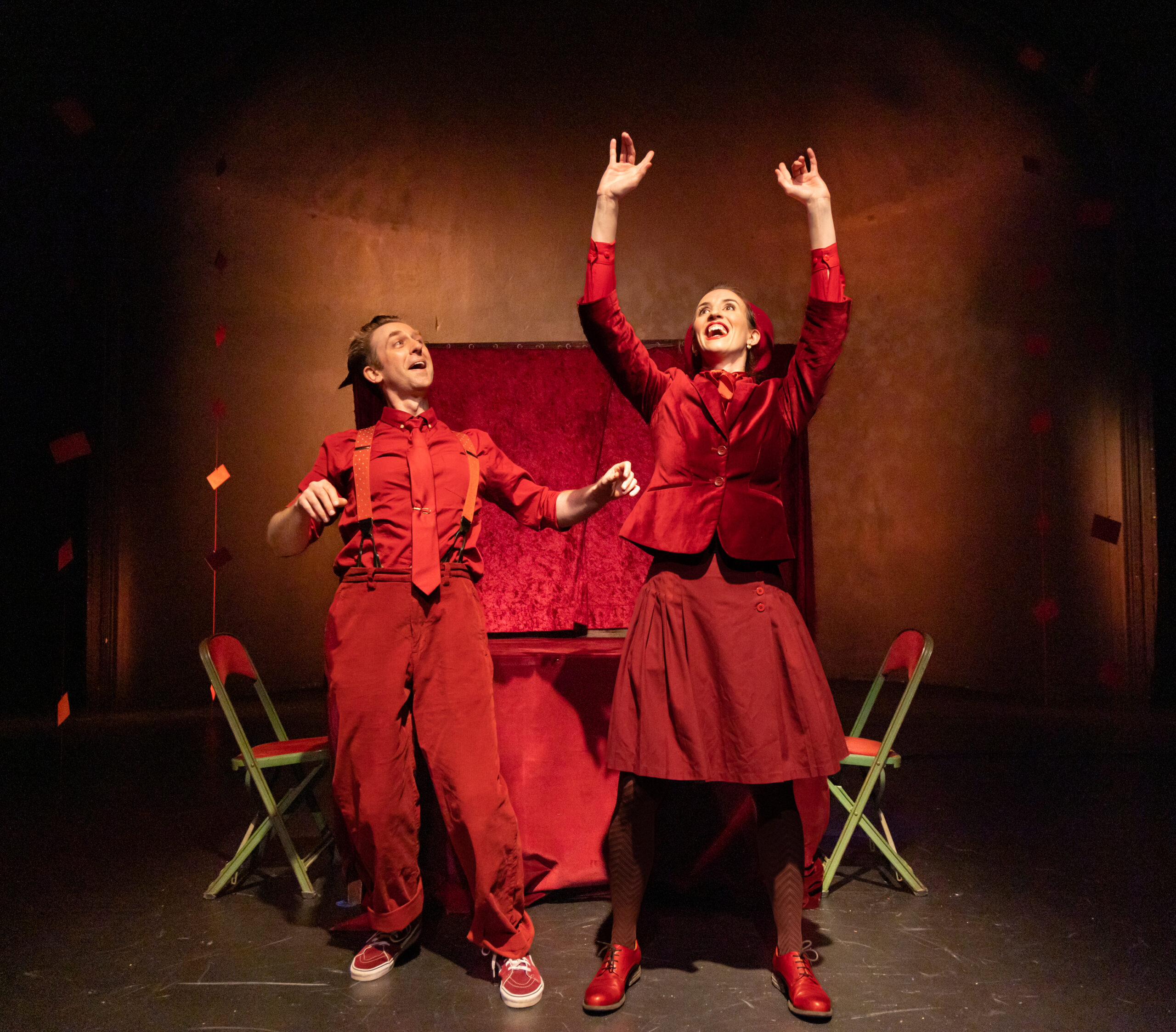 Two performers dressed in red stand on stage with raised arms beside two folding chairs.