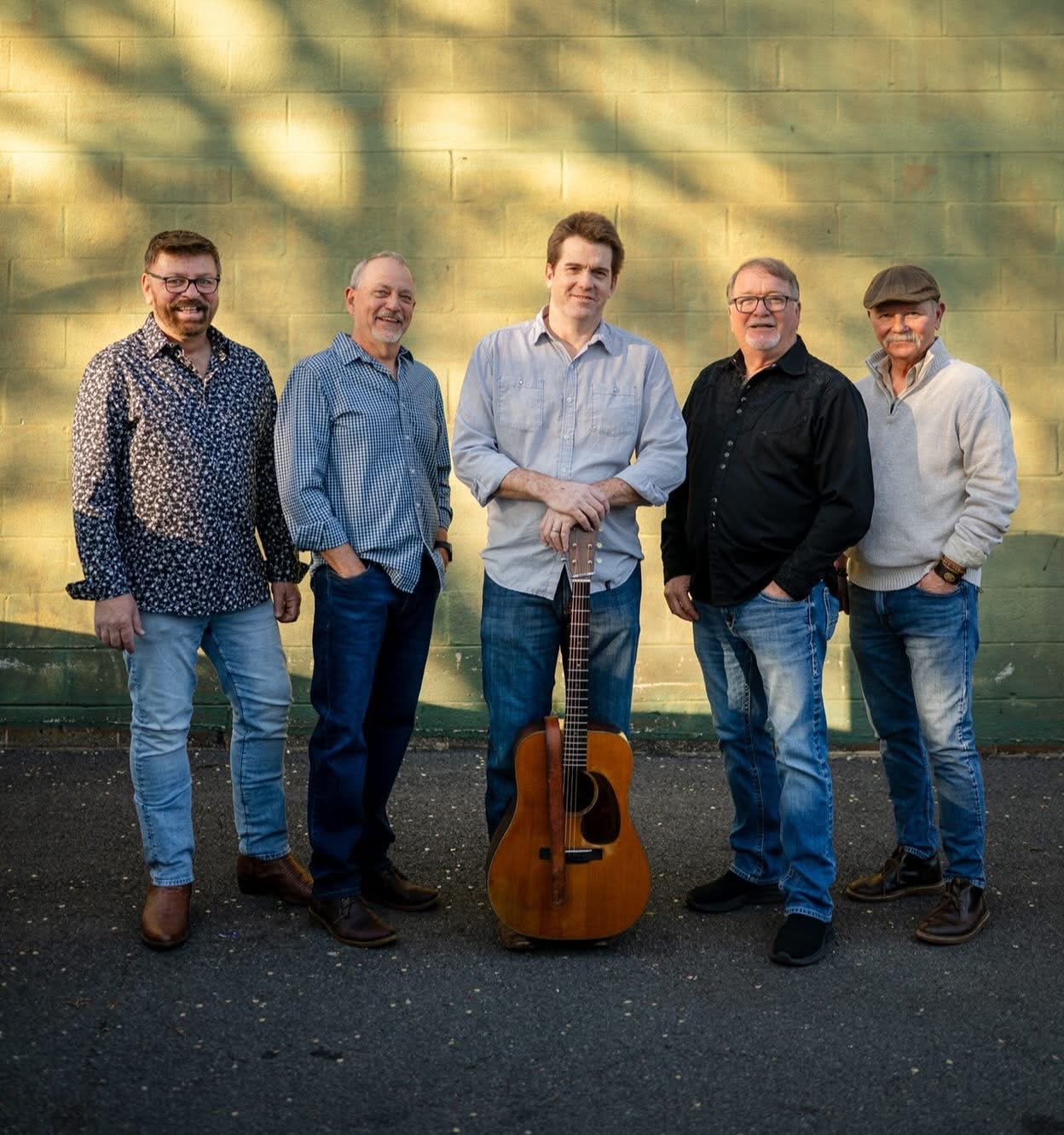 Five members of a folk band stand outdoors against a green brick wall, with an acoustic guitar positioned in front of the center member.