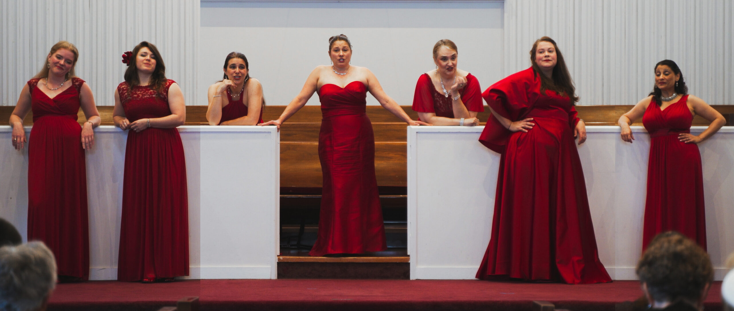 Seven women in red gowns perform together on an indoor stage.