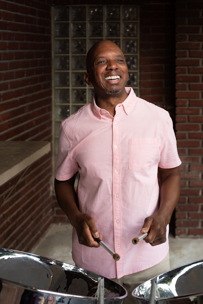 A smiling musician plays steel drums while standing outdoors in front of a brick wall