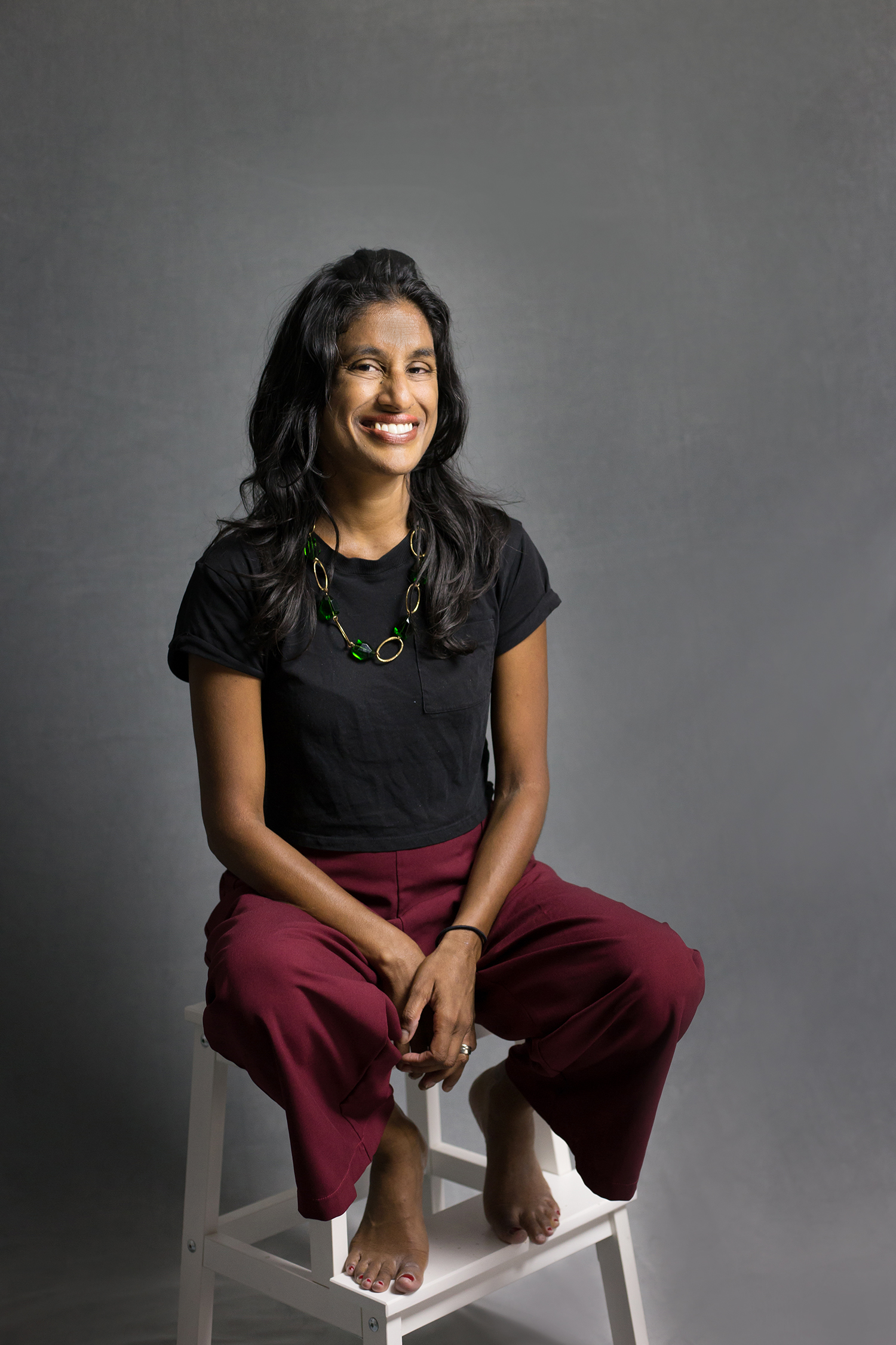 Portrait of author Vara seated on a white stool, smiling against a gray background.