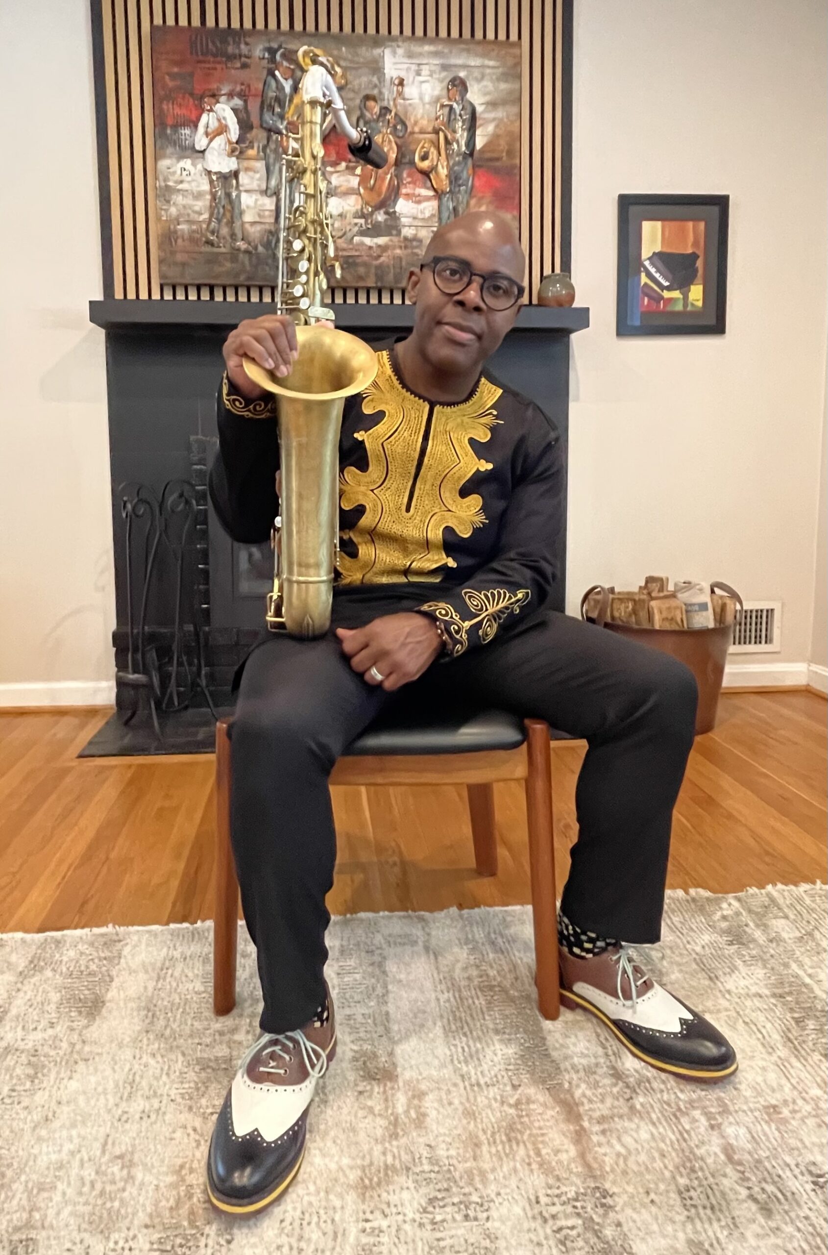 A man in traditional attire sits holding a saxophone in front of a fireplace.
