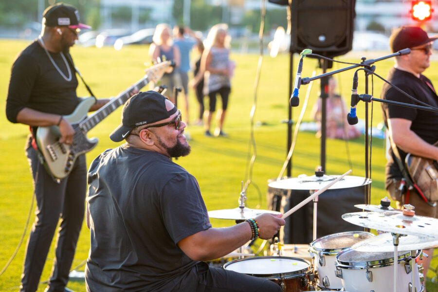 Rear view of a drummer playing a drum set on a grassy field during an outdoor concert, with a bassist and guitarist nearby and a small crowd of people standing and watching in the background.